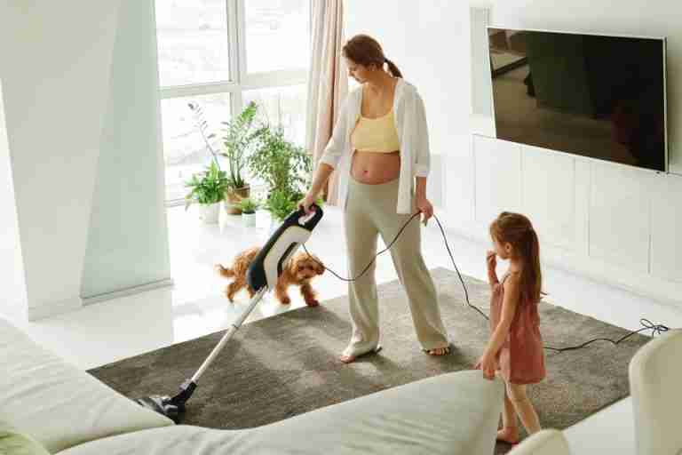 Pregnant mother cleaning carpet with vacuum as daughter and dog play indoors.